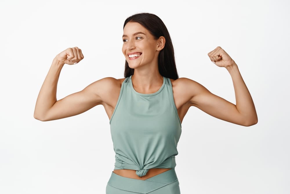 Woman in tank top flexes to show off her toned arms after removing loose skin with brachioplasty